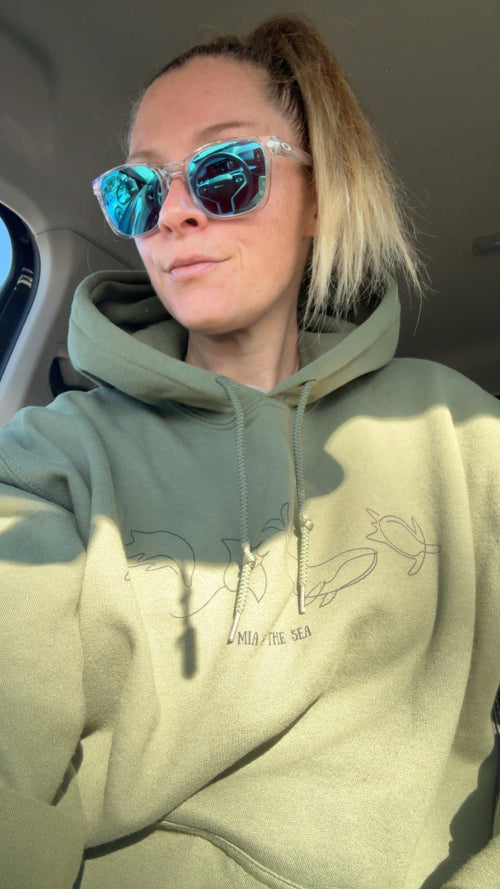 Woman sitting in a car wearing The Marine Hoodie in military green, with dolphin, manta ray, whale and turtle line art on the chest