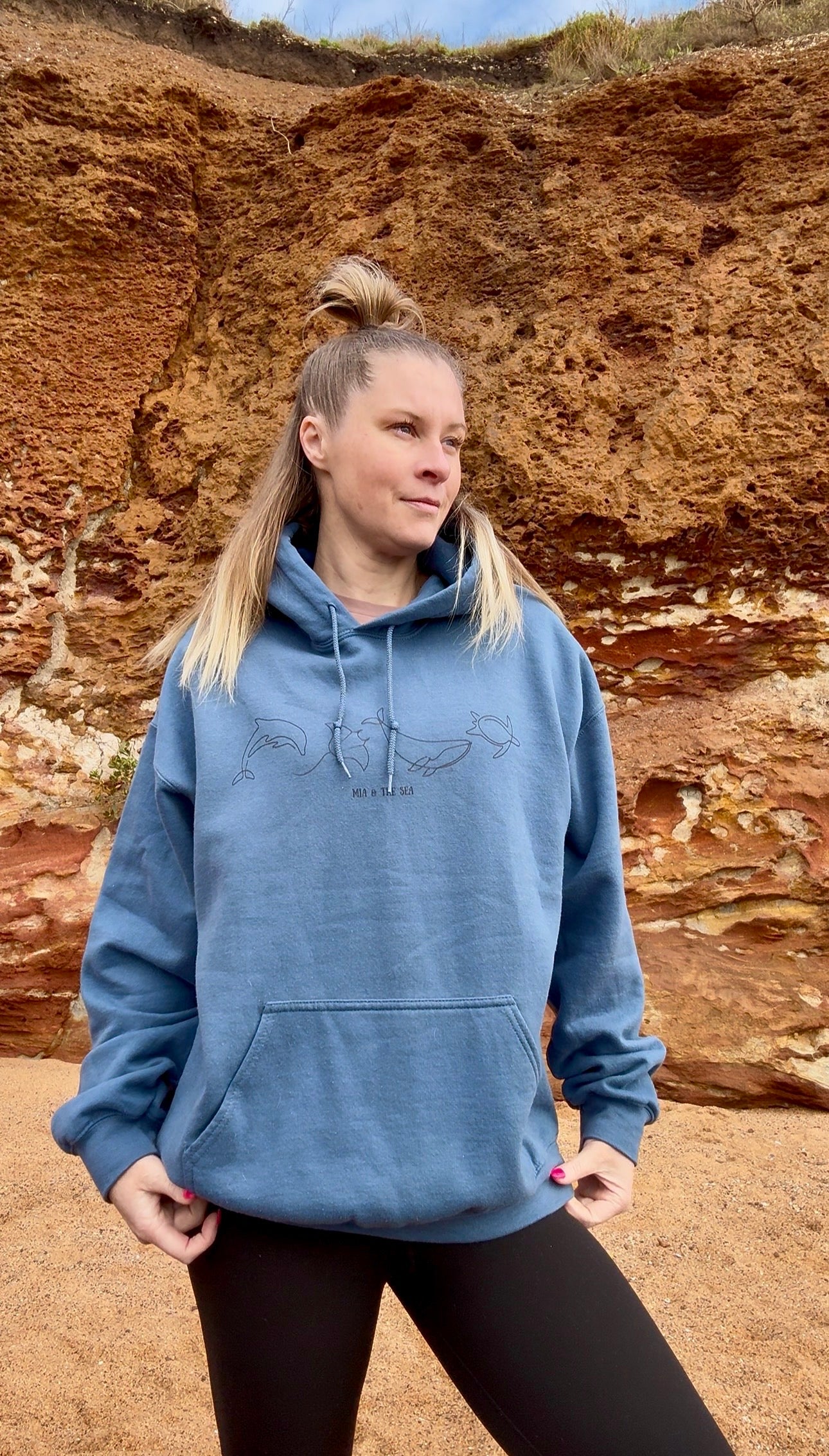 Woman on the beach wearing The Marine Hoodie in indigo blue, with dolphin, manta ray, whale and turtle line art on the chest