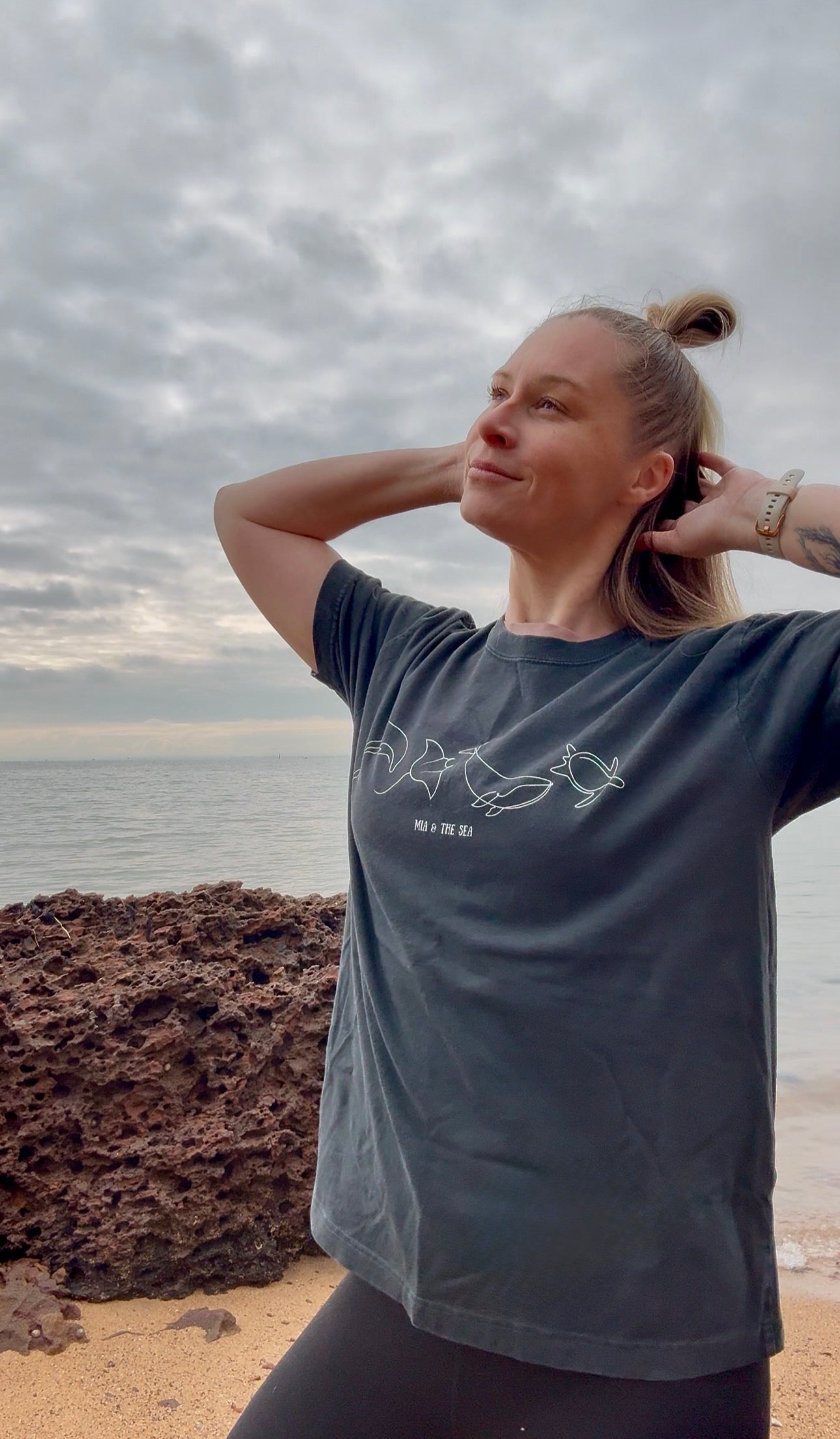 A woman with blonde hair by the ocean under a cloudy sky, wearing the Washed Black Marine Tee