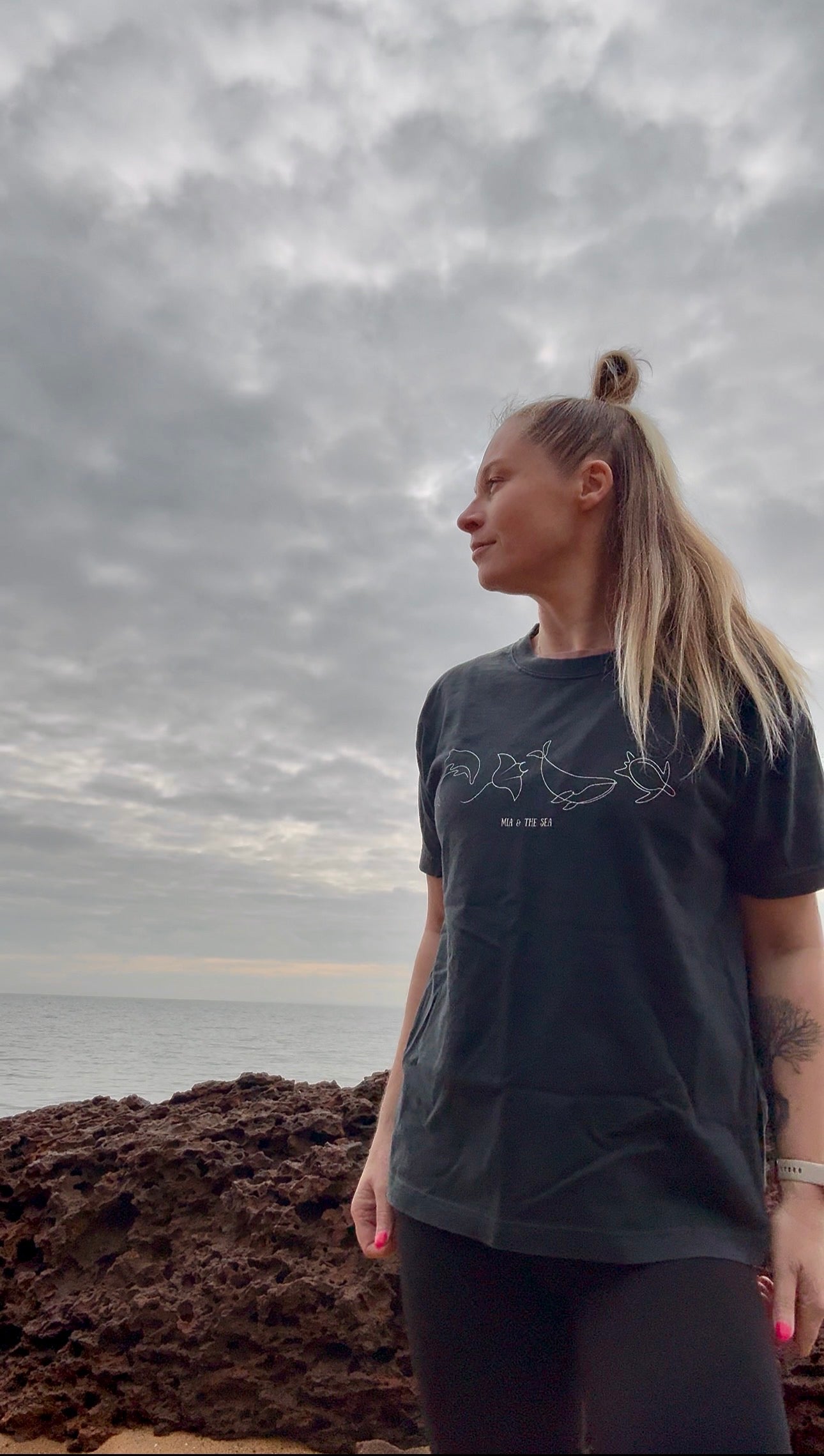 A woman with blonde hair by the ocean under a cloudy sky, wearing the Washed Black Marine Tee