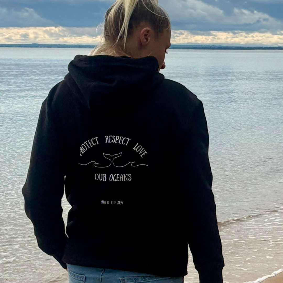 Woman on the beach wearing the Protect. Respect. Love Our Oceans hoodie in black, back view showing the printed design