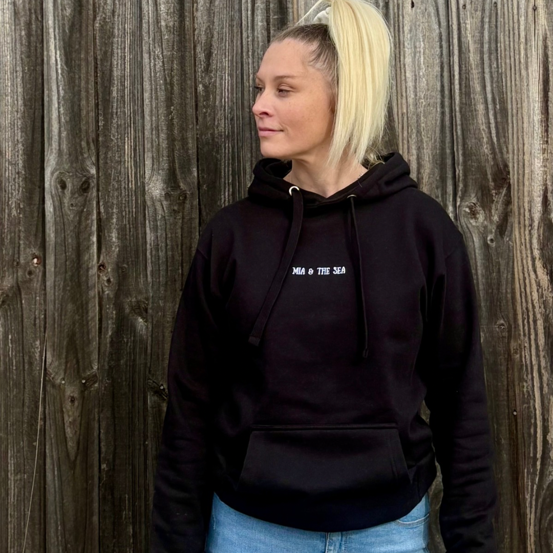 Woman against a fence wearing the Protect. Respect. Love Our Oceans hoodie in black, front view with the embroidered logo