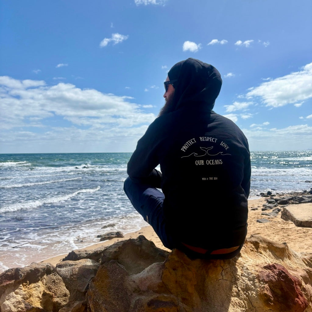 Man sitting on a rock on the beach wearing the Protect. Respect. Love Our Oceans hoodie in black, back view showing the printed design