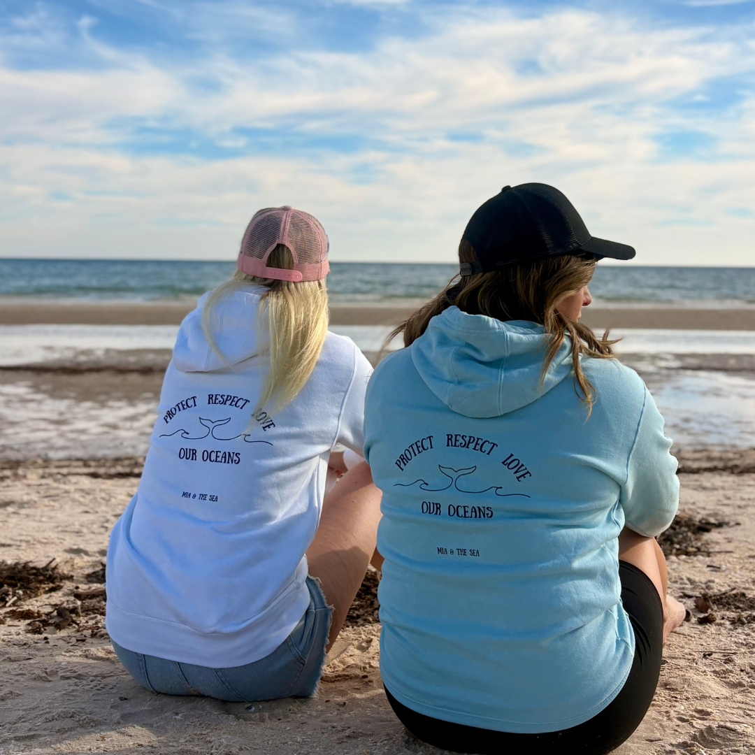 Two women sitting on a beach wearing Protect Respect Love hoodies with ocean conservation messages. Back view.