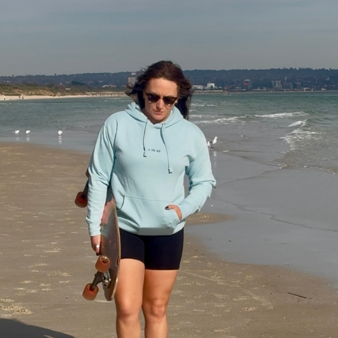 Woman wearing a light blue Protect Respect Love hoodie front view and black shorts walking on a beach with a skateboard.