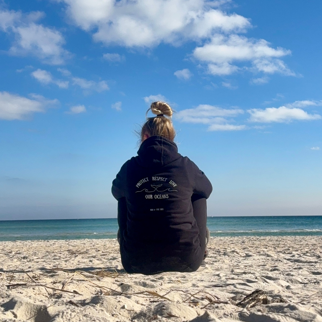 Woman on the beach wearing the Protect. Respect. Love Our Oceans hoodie in black, back view showing the printed design