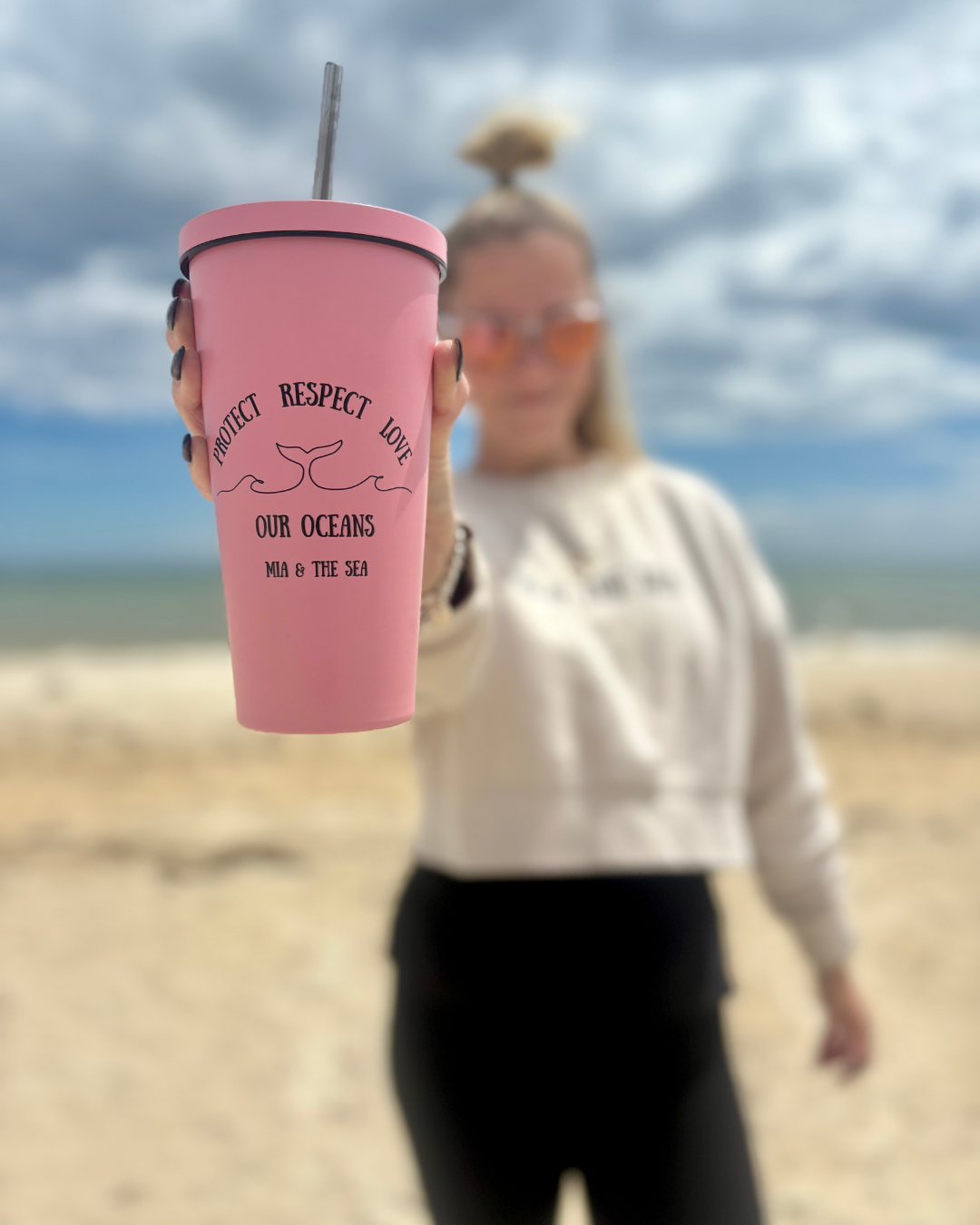 Woman on the beach holding the Ocean Tumbler in pink, with the Protect. Respect. Love Our Oceans logo